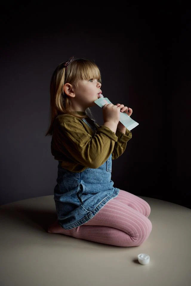 Young girl sitting on a beige surface holding a Haps smoothie bag against a dark background.